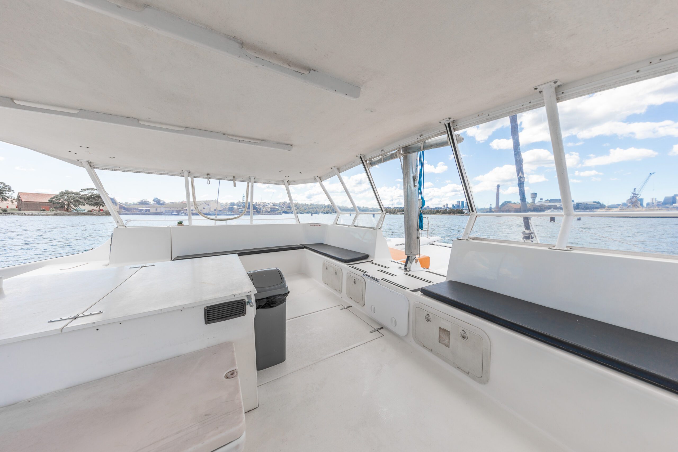 Interior of a white boat with bench seating, large windows showing water and city skyline in the background under a partly cloudy sky.