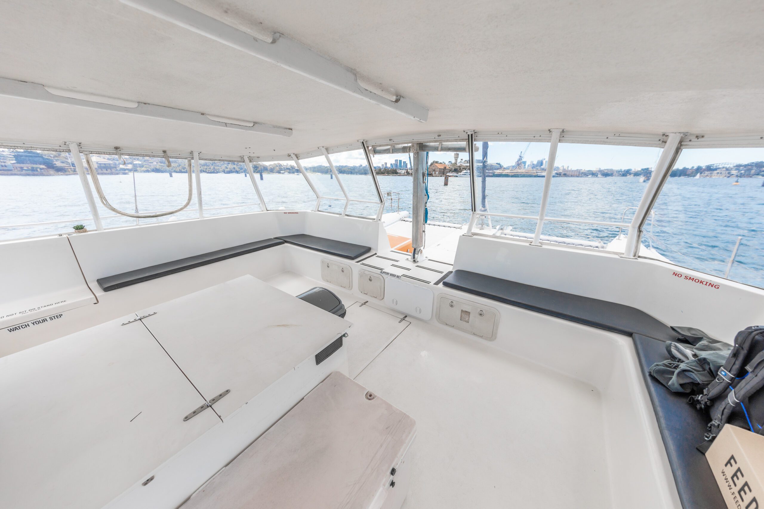 The interior of a white boat with built-in seating, large windows, and views of calm water and a distant city skyline under a bright sky.