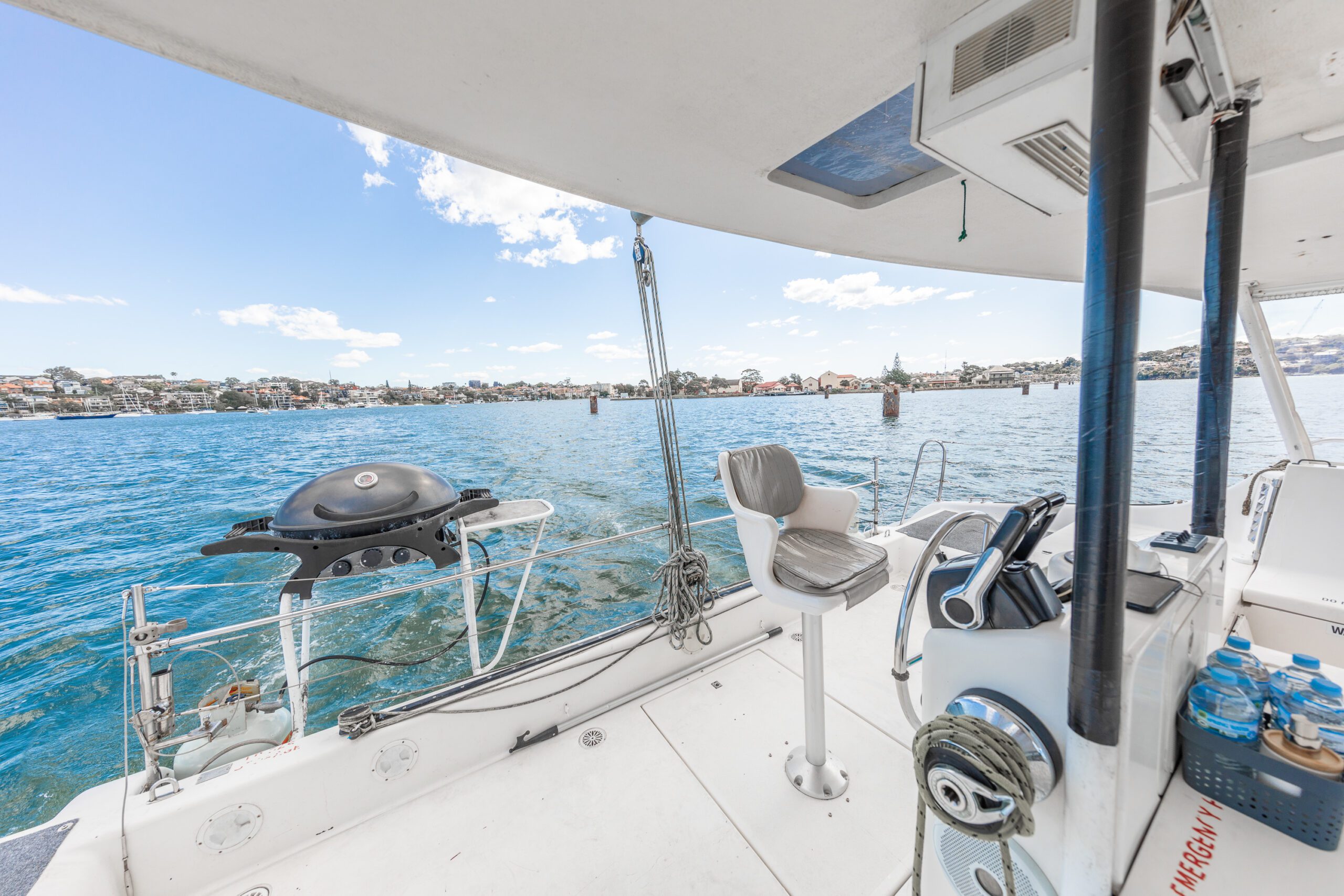View from a boat deck with a grill, chair, and navigation equipment overlooking calm blue water and distant shoreline under a partly cloudy sky.