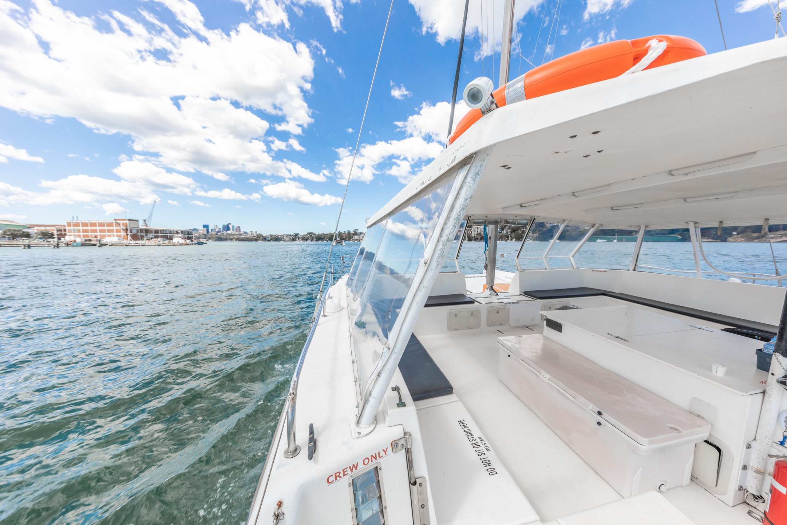 A white boat with a covered seating area sails on blue water under a partly cloudy sky. A city skyline and a stadium are visible in the distance.
